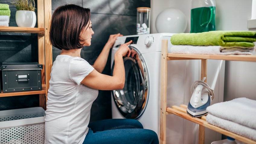 Woman wearing white shirt choosing program on washing machine in laundry room.