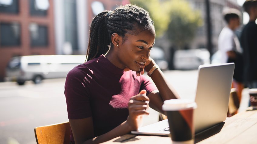 Woman with braids using a laptop at an outdoor cafe.