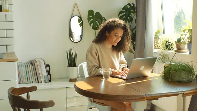 Beautiful young brunette woman sitting in cozy kitchen by a table and working on her laptop computer.