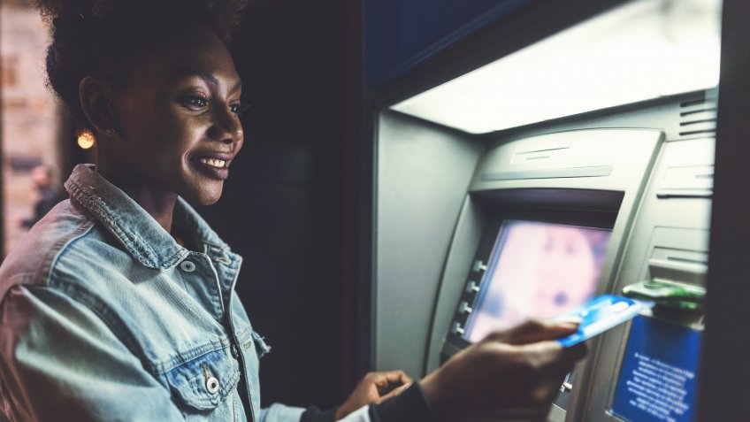 Cheerful African-American woman using her bank card at the ATM in the evening.