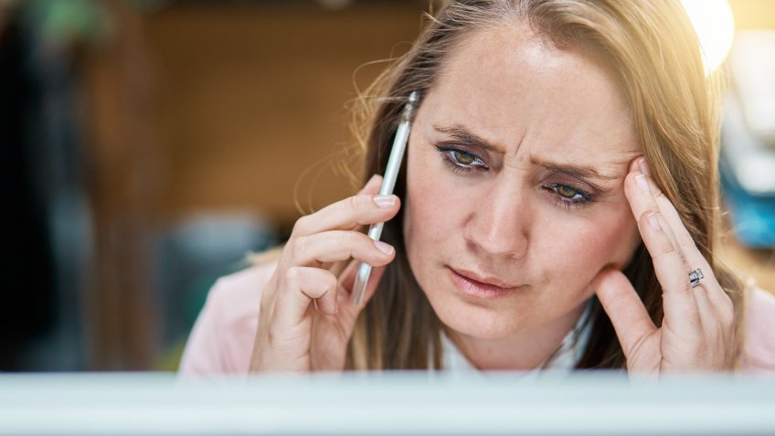 A blonde woman office worker seated in front of a pc monitor frowns, her head on her hand, her other hand holding a mobile phone to her ear.