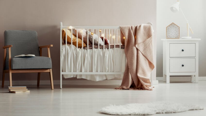 Stylish vintage armchair next to white wooden crib with pastel pink blanket, pillows and cotton ball lights - Image.