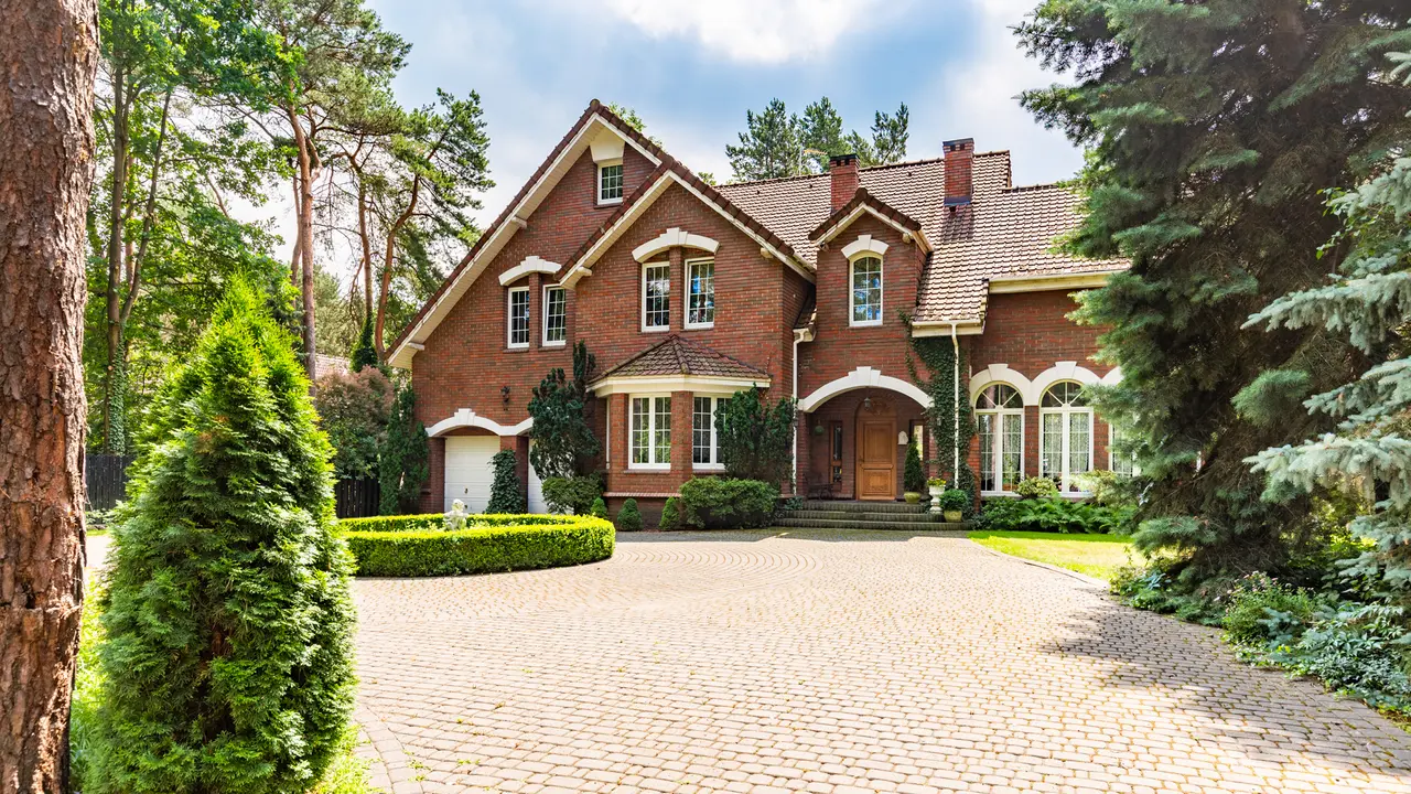 Large cobbled driveway in front of an impressive red brick English design mansion surrounded by old trees.