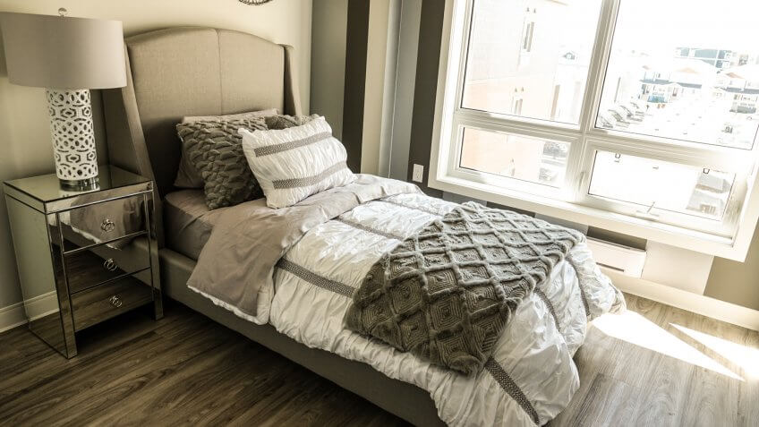 Neutral toned bedroom interior with twin size bed and open window with beautiful hardwood floors and silver mirrored side table.
