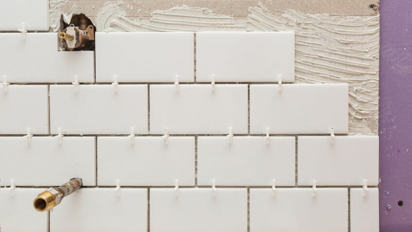White subway tile being installed on backer concrete board with pipes.