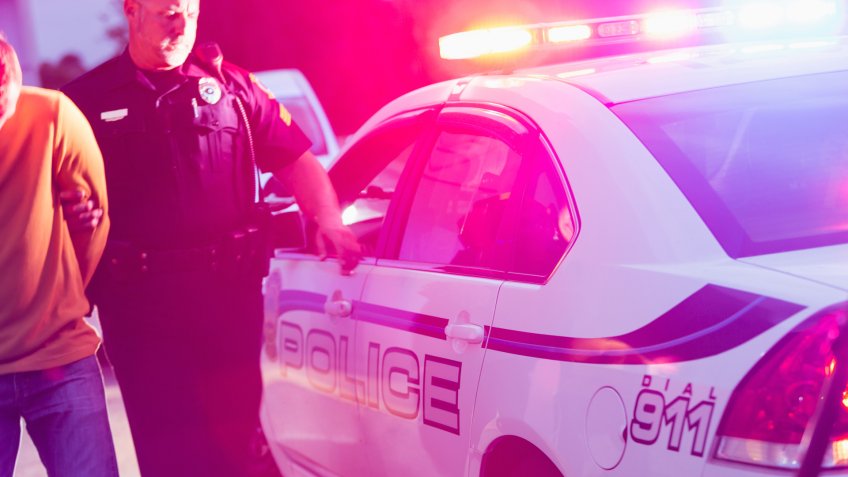 Police officer making an arrest, escorting a young man in handcuffs toward the back seat of his police car, at night.