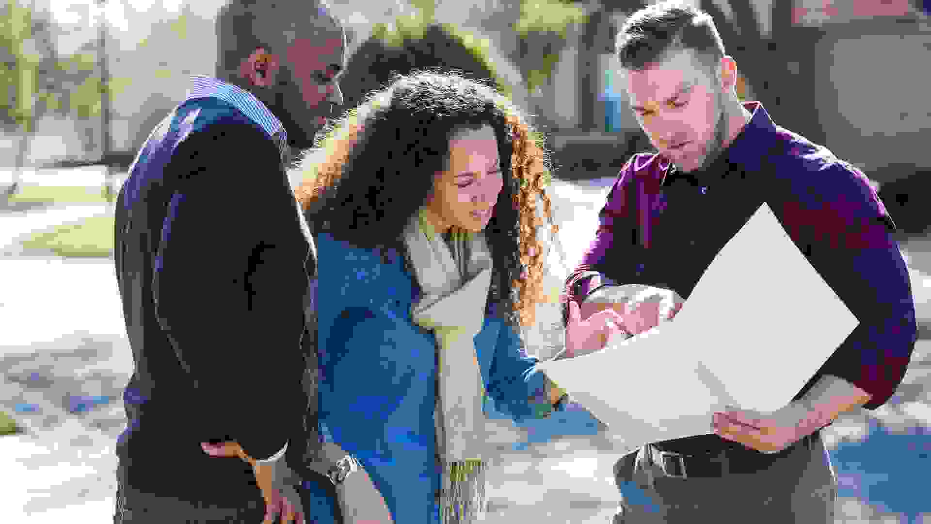 An African American couple standing outdoors on a sunny day on a residential street, with a young man holding a folder.