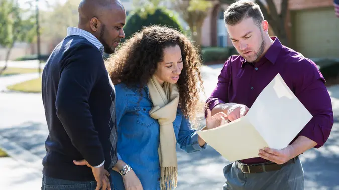 An African American couple standing outdoors on a sunny day on a residential street, with a young man holding a folder.