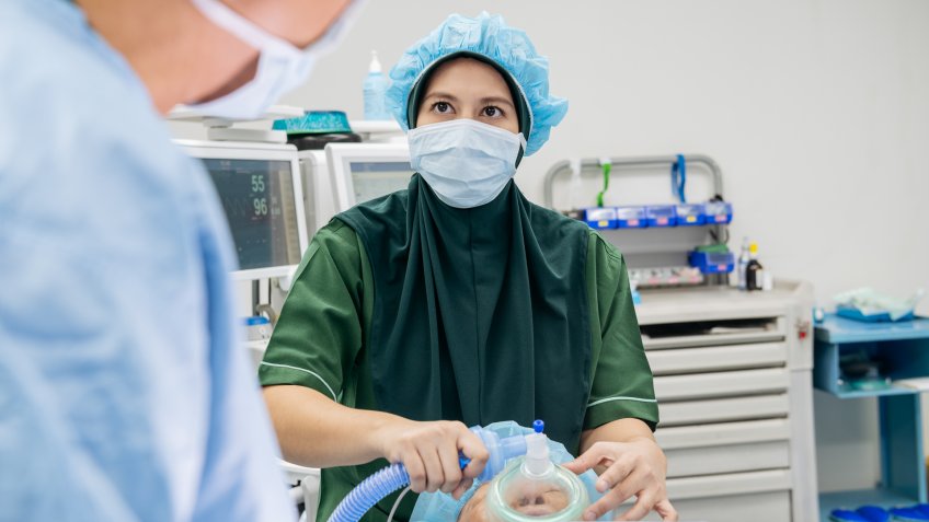 Female anaesthetist holding mask on senior man's face and looking towards colleague with serious expression.
