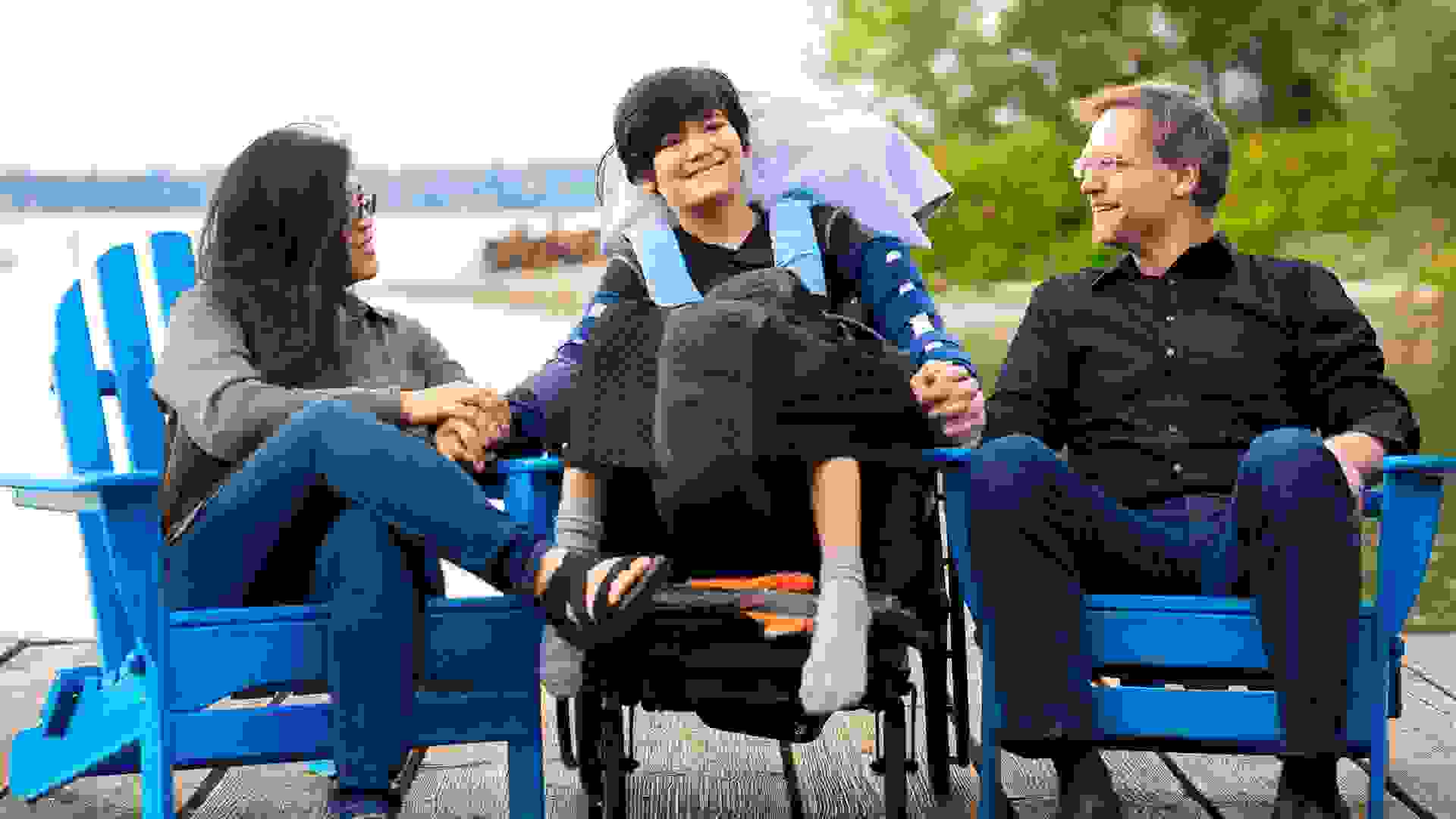 Multiracial couple sitting with disabled twelve year old son in wheelchair while sitting in blue adirondack chairs on wooden pier by lake on summer day.