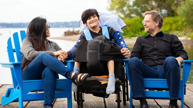 Multiracial couple sitting with disabled twelve year old son in wheelchair sitting in blue adirondack chairs on wooden pier by the lake on summer day.