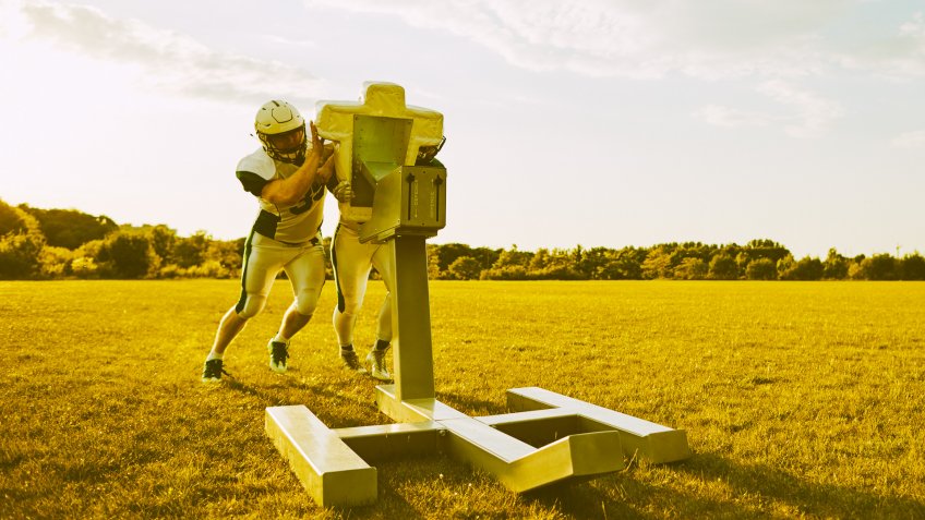 Two American football teammates practicing tackles and defensive drills with a sled outside on a sports field in the afternoon.