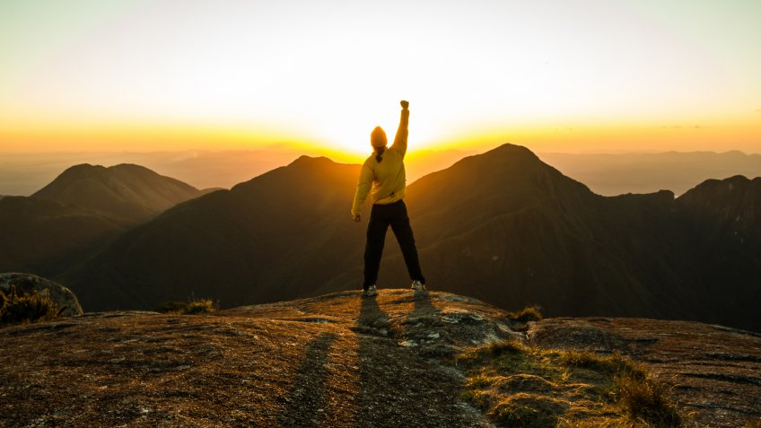Man celebrating success on top of a mountain with one arm Raised and Hands Closed.