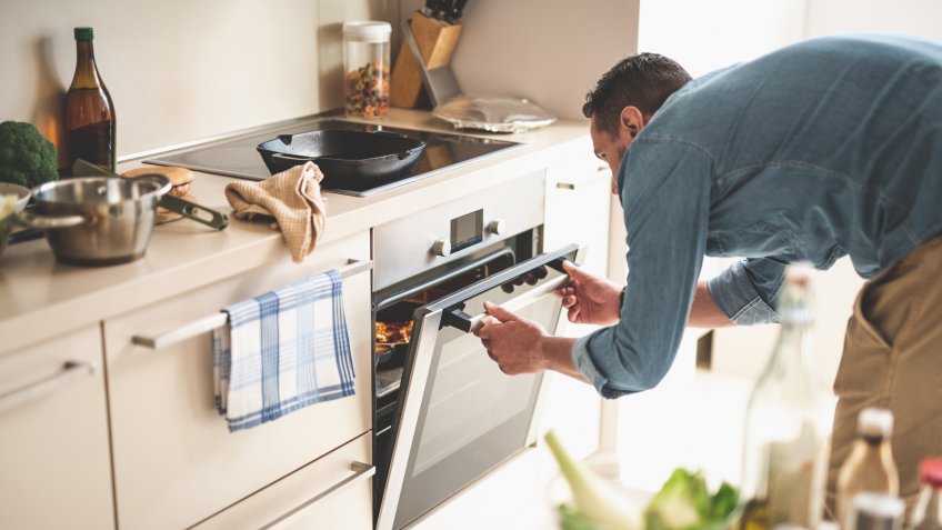 Portrait of gentleman in denim shirt closing door of oven while looking at frying pan with beef steak - Image.