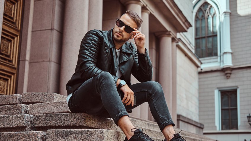 Fashionable guy dressed in a black jacket and jeans holds a smartphone sitting on steps against an old building in Europe.