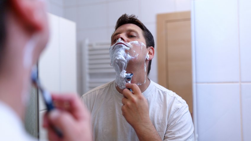 Young gay shaving with razor and shaving cream in bathroombeauty, skin care and people concept - close up of smiling young man applying cream to face and looking to mirror at home bathroom.