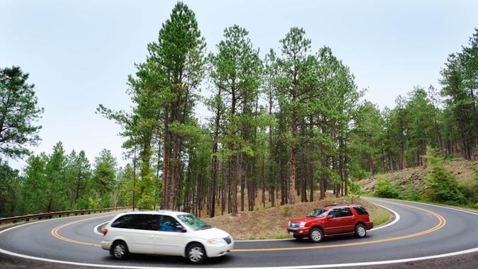 Tourists driving a red car and white sports utility vehicle or SUV around a hairpin curve on a winding road through a scenic forest near Mt.