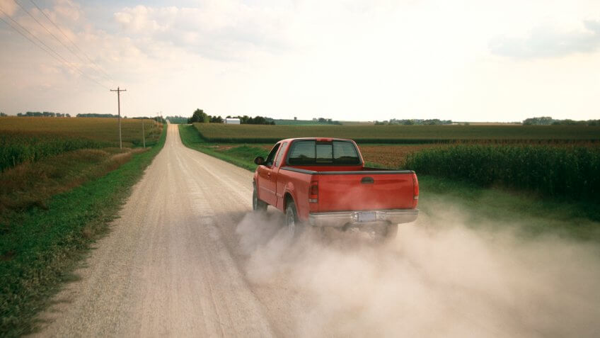 Red pick up truck traveling down dusty rural road.