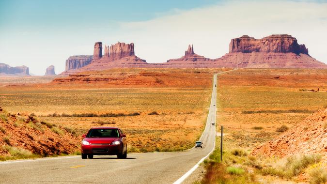 Subject: Tourists in a red car traveling in the American Southwest, driving downhill on a straight length of highway stretching from southern Utah toward Monument Valley, Arizona.