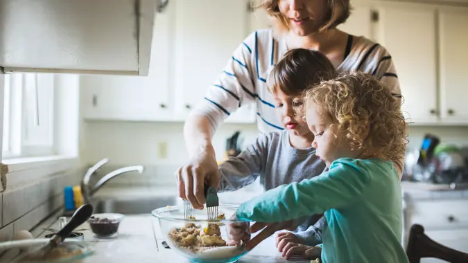 A happy young family prepares to bake chocolate chip cookies together, a boy and a girl about toddler age helping mix and stir the ingredients.