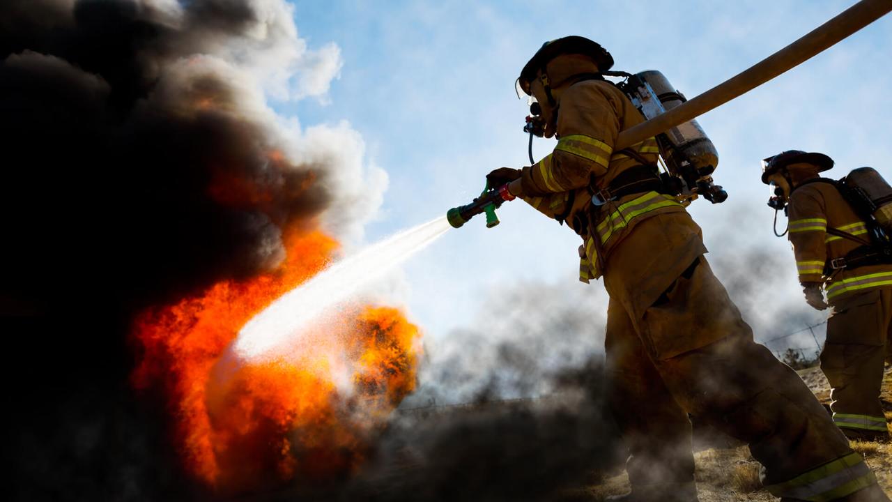 Firefighters in a fire protection suit wearing firefighter helmet with breathing device and holding fire hose is extinguishing a burning house fire that is putting off excessive heat and smoke.