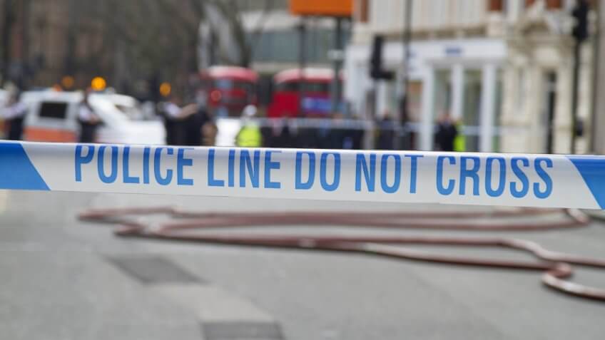 Police Line Do Not Cross tape against blurred city background,police van and fire engine hoses visible in distance.