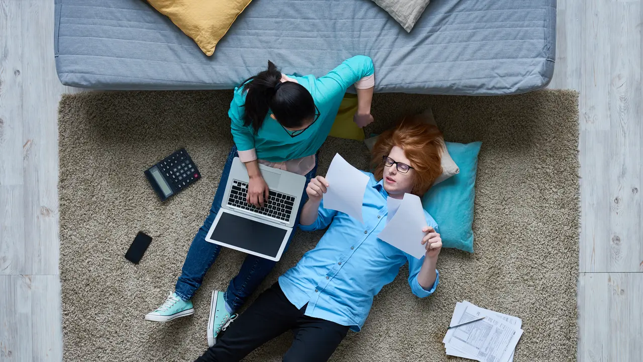 Tired redhead guy lying on carpet and reading tax documents while his girlfriend listening to him and using laptop.