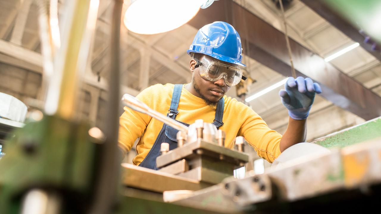 Serious busy young black factory engineer in hardhat and safety goggles examining milling lathe and repairing it while working at production plant.