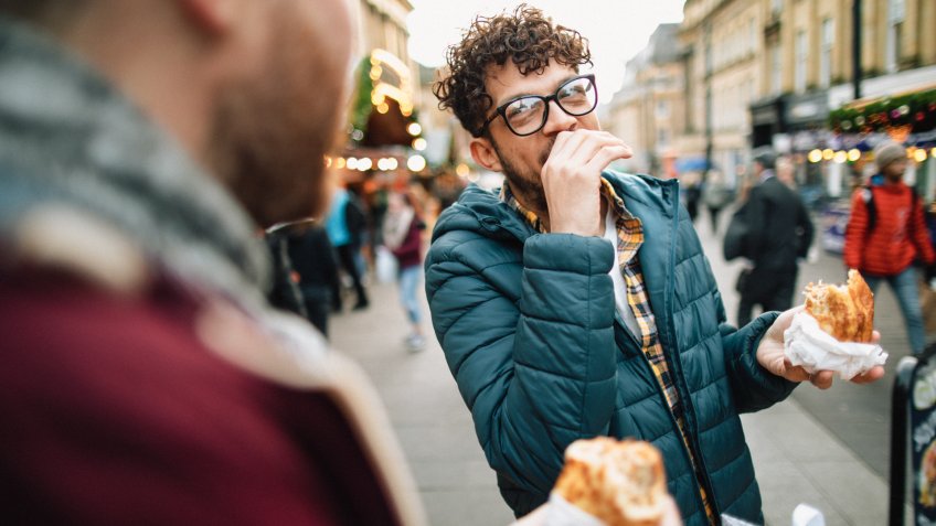 Two young gay men are eating hot food at a Christmas market.