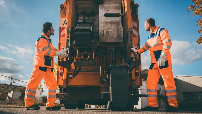 Two refuse collection workers loading garbage into waste truck emptying containers.