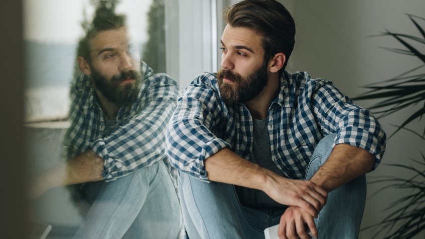 Young pensive man sitting by the window sill and looking through it.