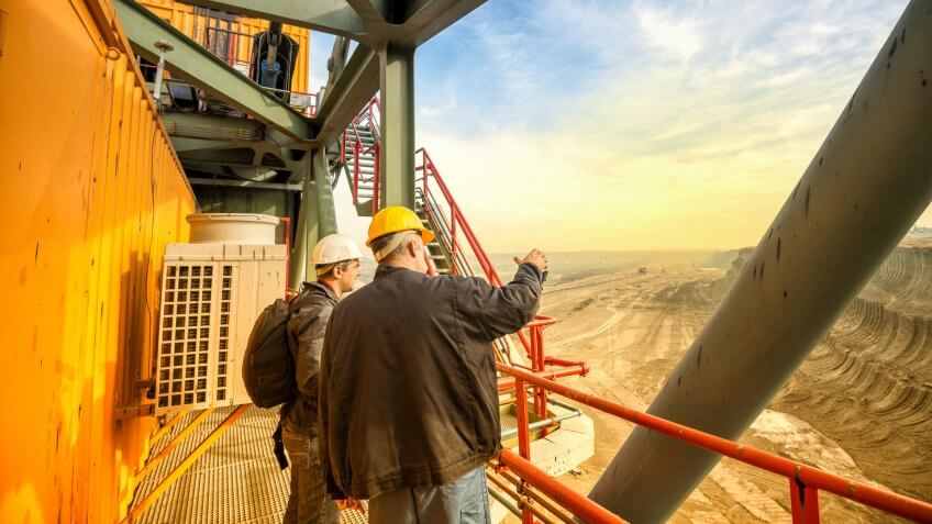 Two coal mine engineers with protective helmets standing on a huge drill machine, chatting and watching at the digging site.