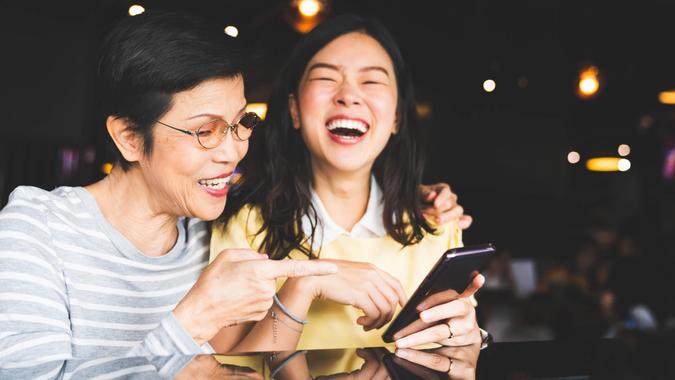 Asian mother and daughter laughing and smiling on a selfie or photo album, using smartphone together at restaurant or cafe, with copy space.