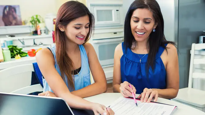 Mother and teenage daughter are in the kitchen researching colleges and filling out university applications.