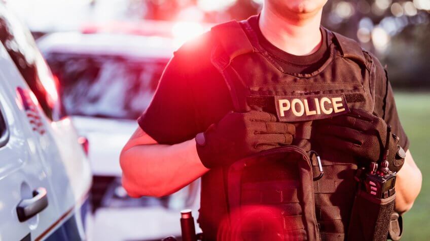 Cropped view of an Hispanic police officer wearing a bulletproof vest, standing beside two police cars.