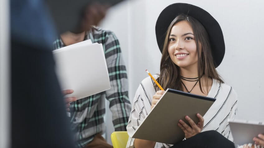 Close up of a young, confident Latino woman, stylishly dressed in a black hat, smiling as she takes notes on a paper pad in a meeting.