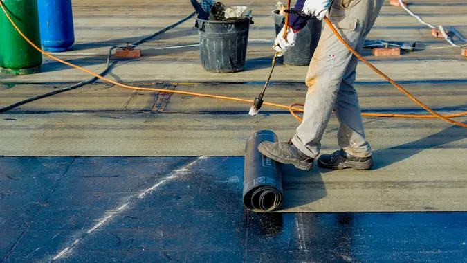Worker preparing part of bitumen roofing felt roll for melting by gas heater torch flame.