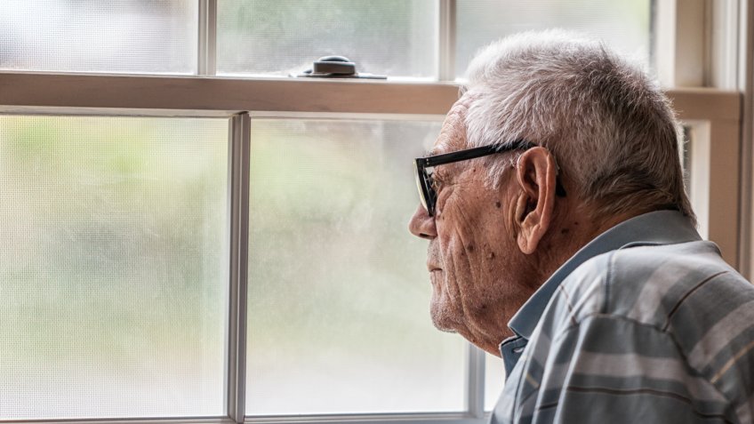 A hearing impaired elderly senior adult man wearing a hearing aid is sitting staring through the hazy, speckled, unwashed window and grungy mesh screen of a living room window at home.