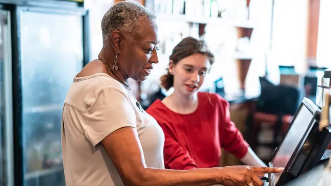 The active senior, 77-years-old, African-American businesswoman, business owner, teaching the new employee, the 18-years-old Caucasian white girl, how to use the computerized cash register in the small local restaurant.