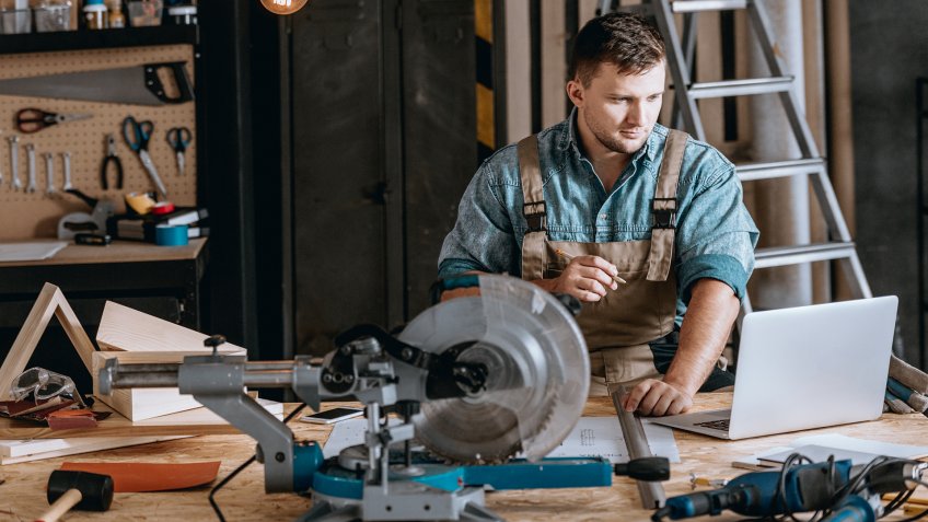 Young bearded carpenter focused on planning woodworking project using laptop in workshop.