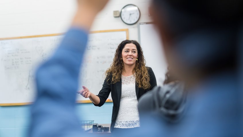A teacher stands up front and calls on one of her students to respond to a question under discussion.