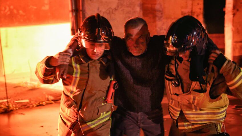 Two firefighters rescuing a victim of a fire with arms around them and helping him to walk away from the fire.