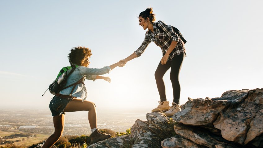 Young woman helping friend to climb up the rock.