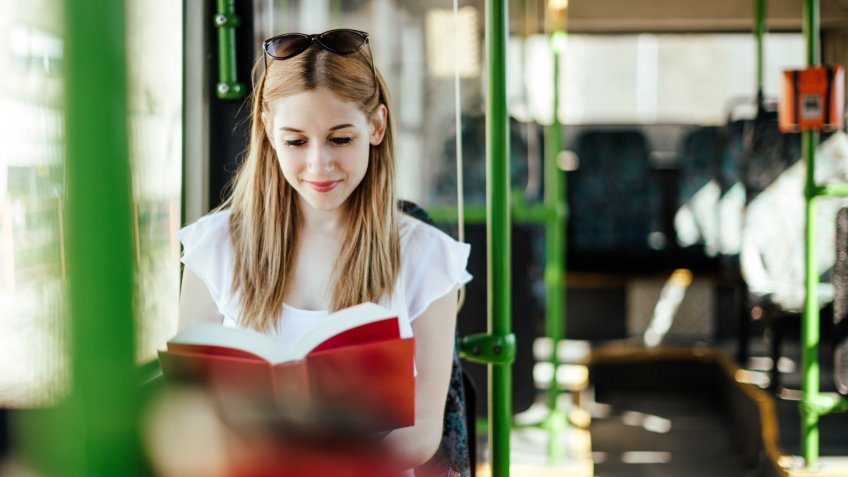 Blonde student girl is reading a book while commuting.