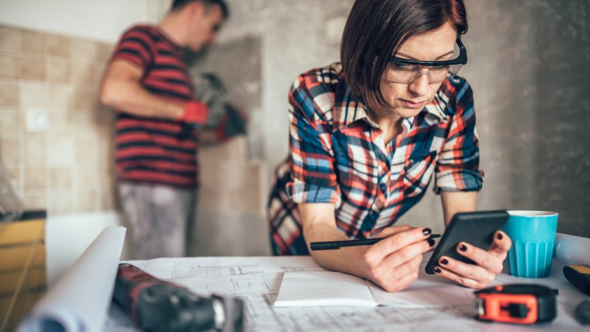 Woman using smart phone on the table while her husband removing old wall tiles in the kitchen.