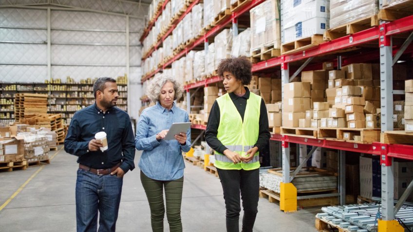 Senior manager using digital tablet while walking in warehouse with supervisor and foreman.