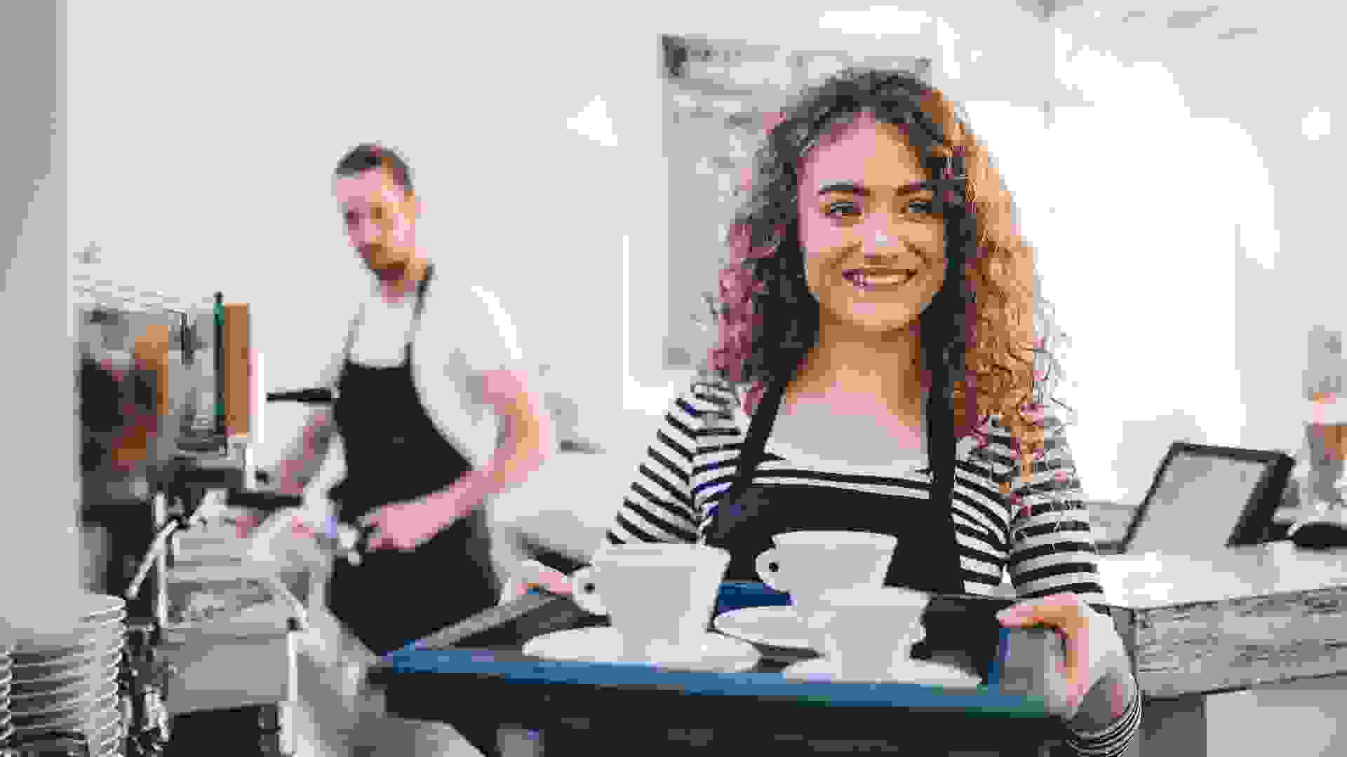 Young beautiful girl carrying cups of coffee on a tray and her colleague operating espresso machine in a coffee shop.