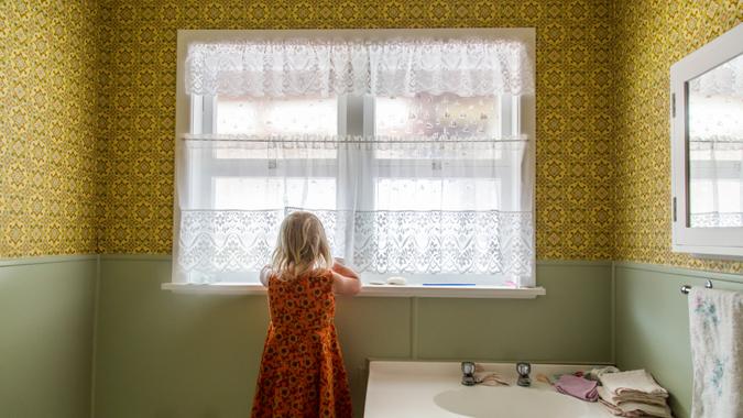 1970s bathroom child looking out window shutterstock_1399745975 Young girl alone exploring the bathroom of a 1970s house.