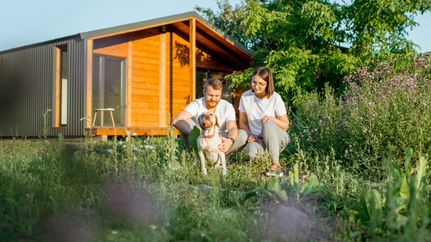 Young couple playing with dog on the backyard of the wooden country house during the sunset.