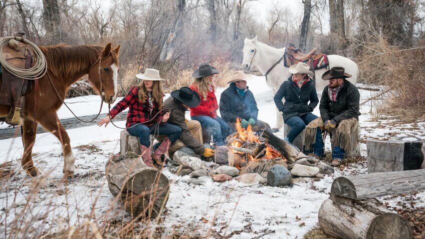 Three Caucasian couples in their 20's huddle around a campfire outdoors with two horses on a winter day, Livingston, Montana, USA.